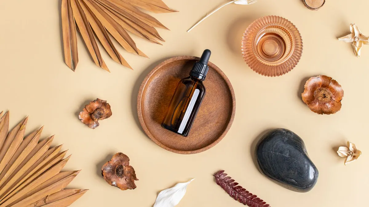 An unbranded amber glass dropper bottle on a round wooden dish, surrounded by dried palm leaves, botanicals, and a smooth stone on a warm beige background