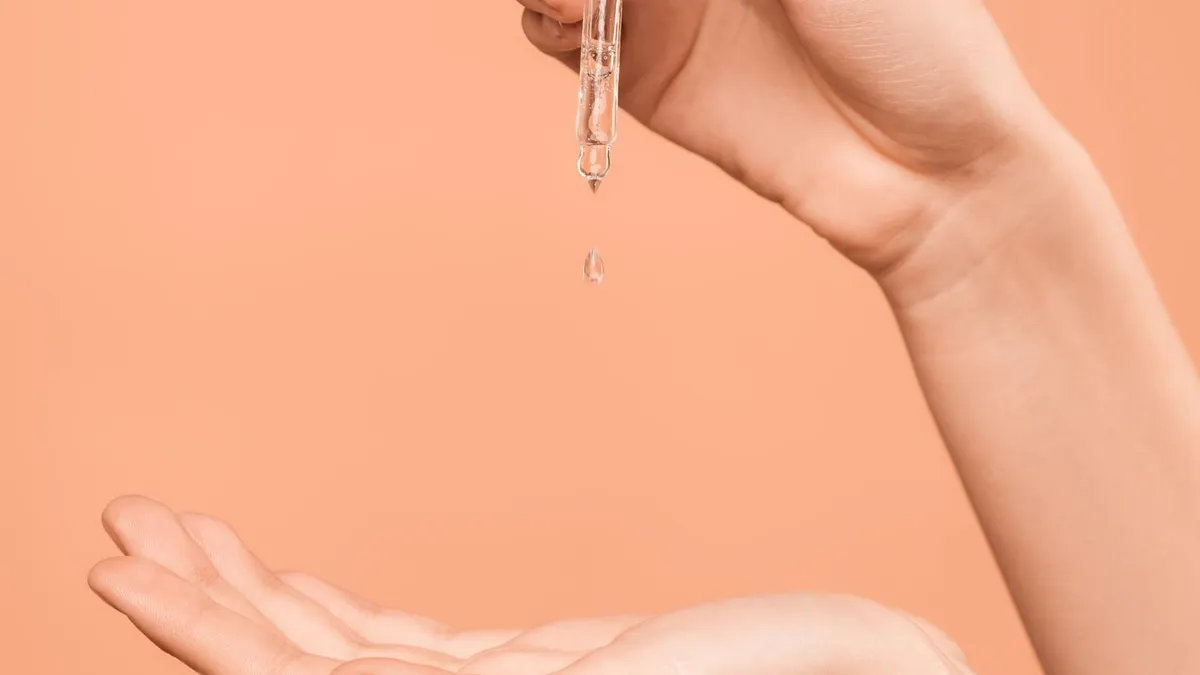 A clear glass dropper releasing a serum drop toward an open palm against a warm apricot background