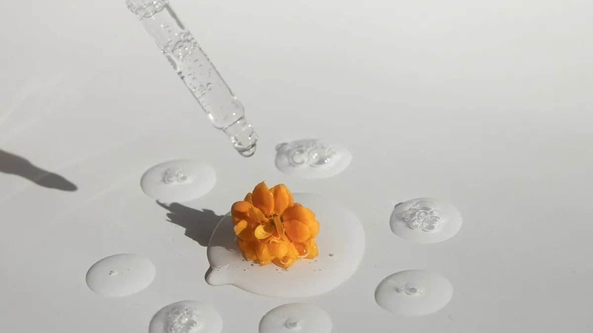 A glass dropper and serum droplets around a single orange flower on a white surface