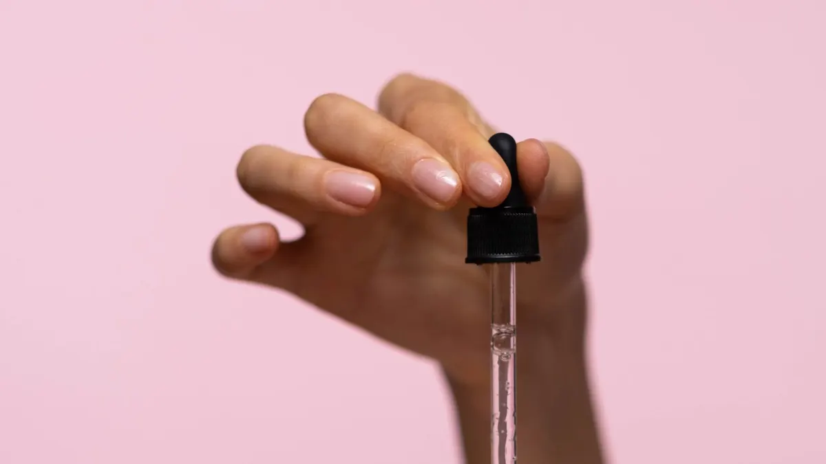 A hand holding a glass serum dropper against a soft pink background